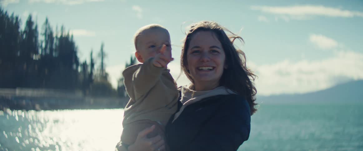 A woman is smiling brightly while holding a baby who is looking and pointing offcamera they are standing outdoors by a large body of water that is sparkling in the bright sunshine