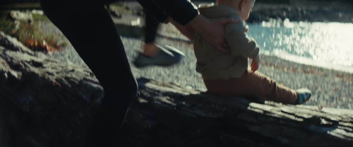 The video begins with an adult helping a toddler climb up onto a large smooth log near the edge of a bright shimmering body of waterthe adult gently lifts the child and then sits down next to them smiling down at the child as they enjoy the sunny ou