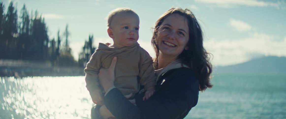 Young mother holding her baby boy son on her arms and kissing him while smiling next to the ocean outdoors during day time