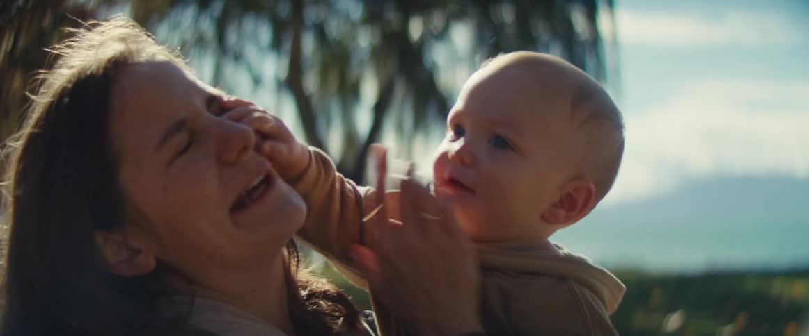 A mother holds her smiling baby outdoors in a sunny park