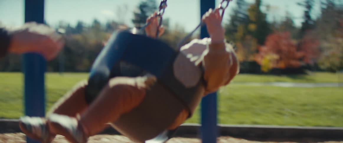 A cheerful baby in a light brown outfit is happily swinging in a baby bucket seat at a park playground an adults arm and hands are visible repeatedly pushing the childs swing on a bright sunny day