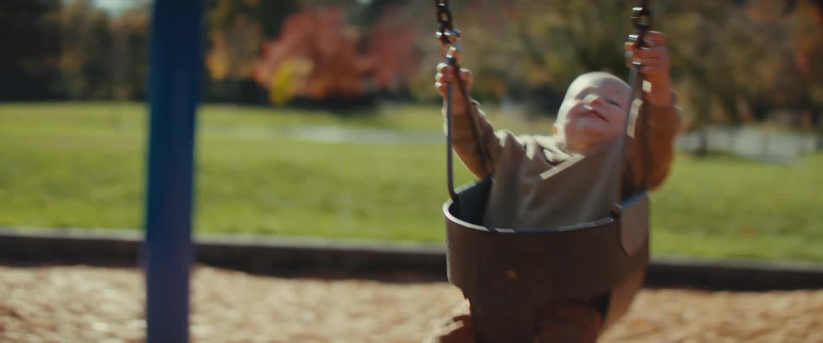 The video features a baby or toddler sitting in a bucket swing at a sunny park with a delighted expression on their face as they swing highan adults hands are visible pushing the swing with a backdrop of a grassy field and trees beginning to show au