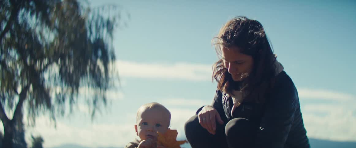 A baby sits on grass nibbling a yellow leaf then crawls away while an adult crouches nearby on a sunny autumn day