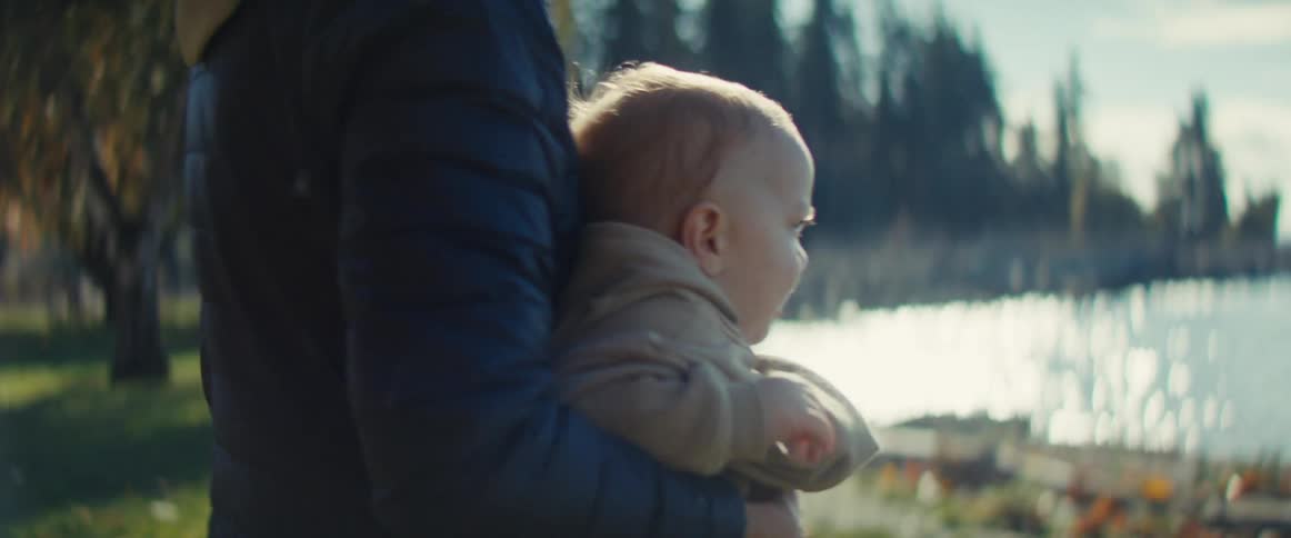Adult carries a baby near a peaceful lakeside with sun reflecting on water