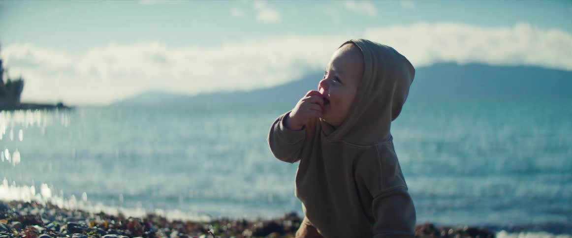 Male baby wearing a brown hoodie crawling on the floor on the coast next to the ocean during day time