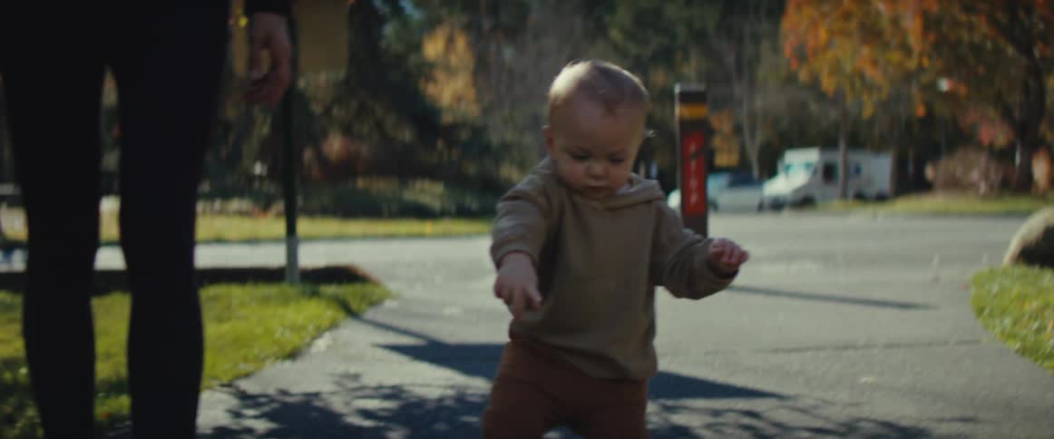 A baby holding an adults hand is walking on a concrete path in a park then lets go of the adults hand and starts walking alone