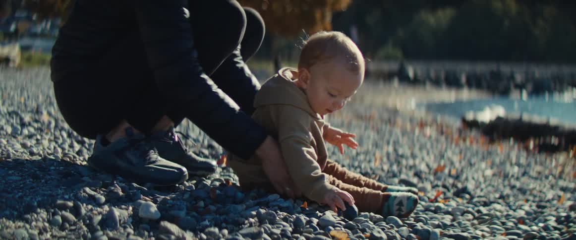 The video shows a closeup of a baby or young toddler in a tan outfit playing on a rocky or pebbled beach or lakeshore on a bright sunny dayan adult wearing dark clothing and sneakers kneels nearby gently watching and interacting as the child picks u