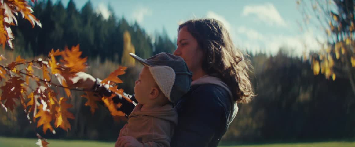A mother holds her toddler son while he plays with autumn leaves on a tree branch