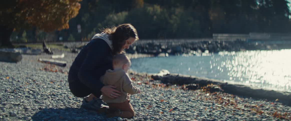 A mother and her baby are walking on a rocky beach when the baby falls prompting the mother to crouch down and hug the child the mother then helps the child stand and they begin to toss small pebbles toward the waters edge
