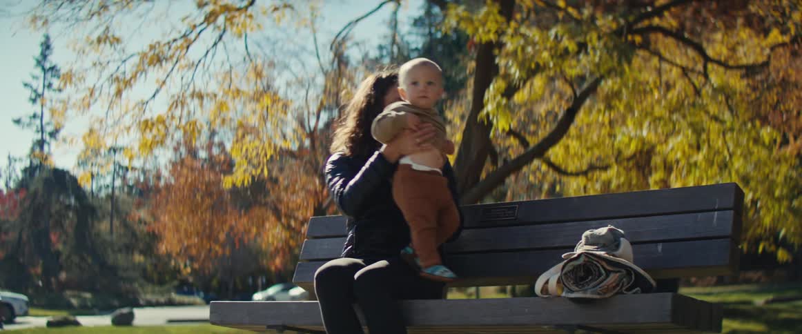 A woman sits on a park bench with her baby surrounded by autumn trees and a peaceful sunny day