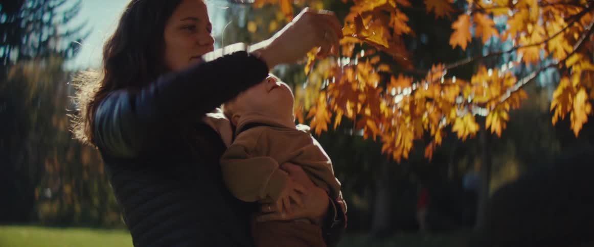A mother holds her baby close on a bright sunny autumn day smiling warmly as they both interact with the vibrant orange and yellow leaves of a nearby tree with the baby curiously reaching for and touching the foliage
