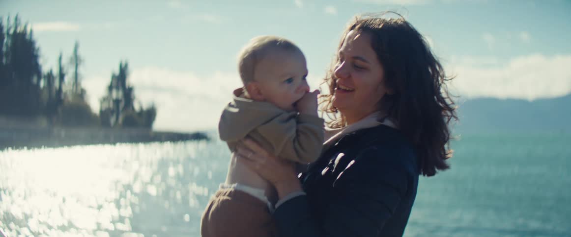 A mother lifts her smiling baby by a sunlit lake surrounded by trees and mountains