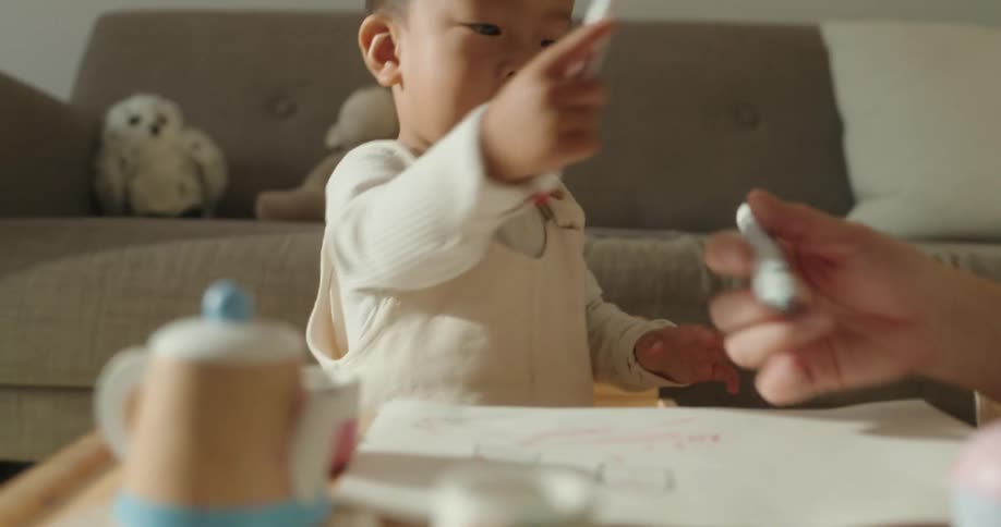 A little boy wearing white clothes is playing with a pen and paper that are in front of him then passing them to an adult
