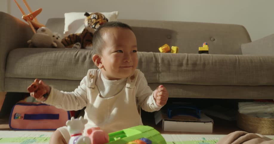 The video features a toddler sitting on the floor in a bright living room joyfully playing with a colorful toy that has a row of brightly colored keys in the background a neutraltoned sofa is visible scattered with several stuffed animals and some bl