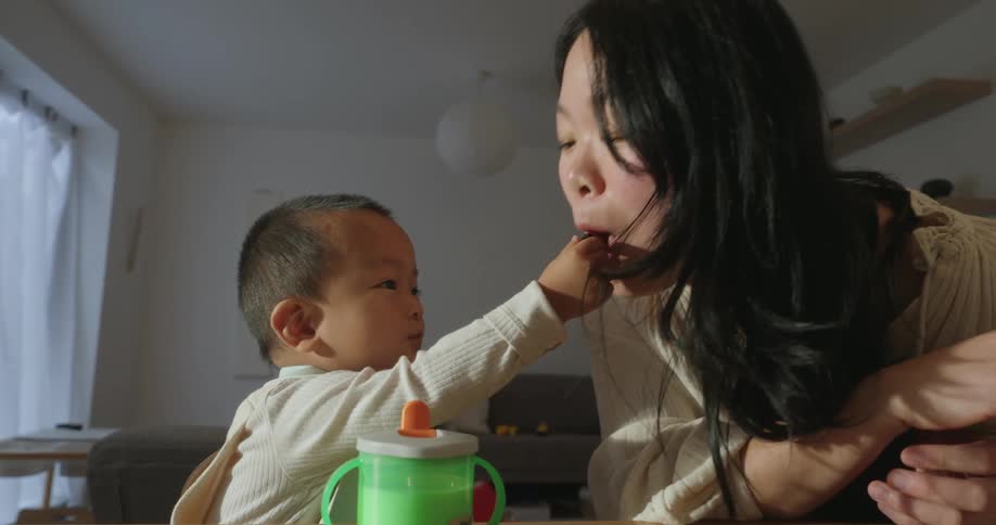 An asian baby playfully feeding his mother while sitting at a table in a living room