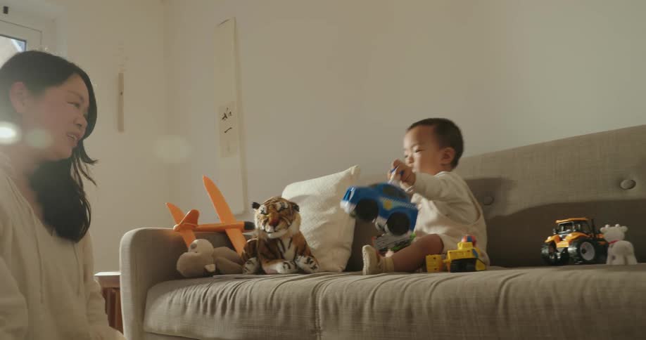 A mother and her baby are playing together indoors with the baby sitting on a couch among several toys including a stuffed tiger and toy vehicles while the mother leans in from the side to interact with the child and the toys