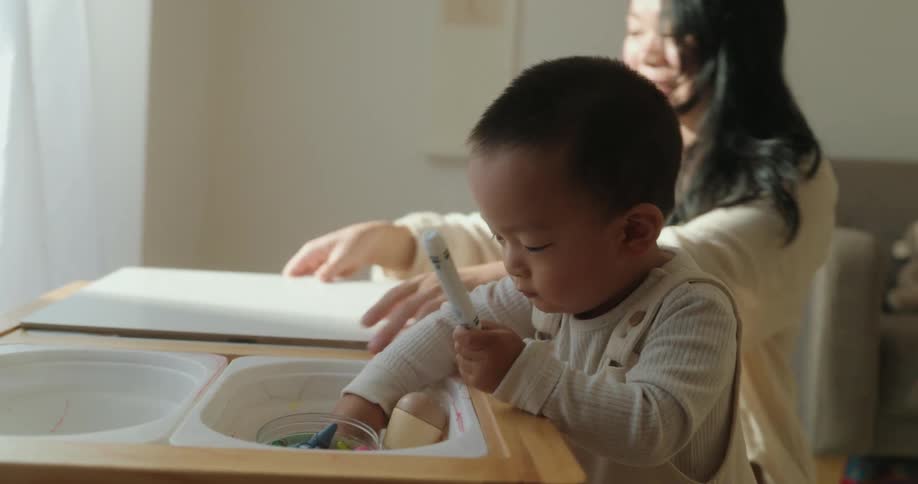 A small child stands at a low wooden table with storage bins taking out a marker to prepare for drawingan adult is seated behind the child placing a large white drawing board onto the tabletop