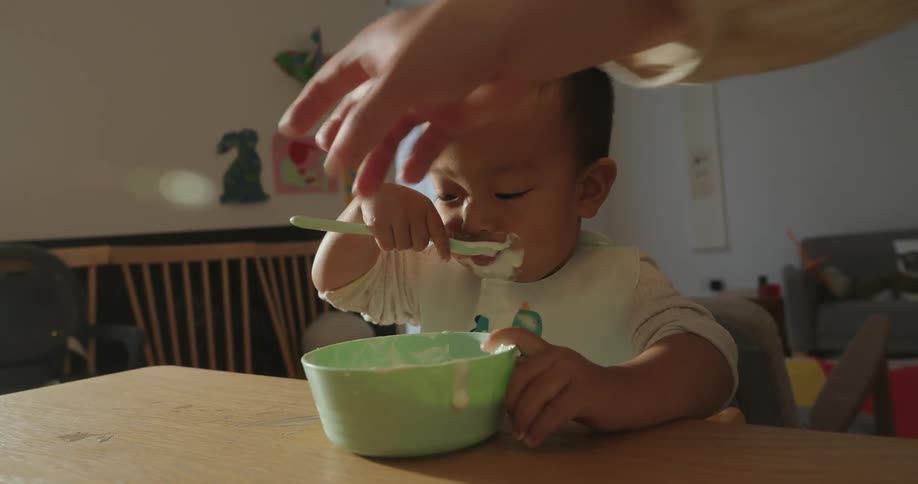 A toddler in a bib eats from a green bowl with a spoon as an adult hand helps guide the bite