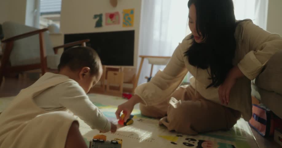 A woman plays on the floor with a baby moving toy cars across a bright play mat