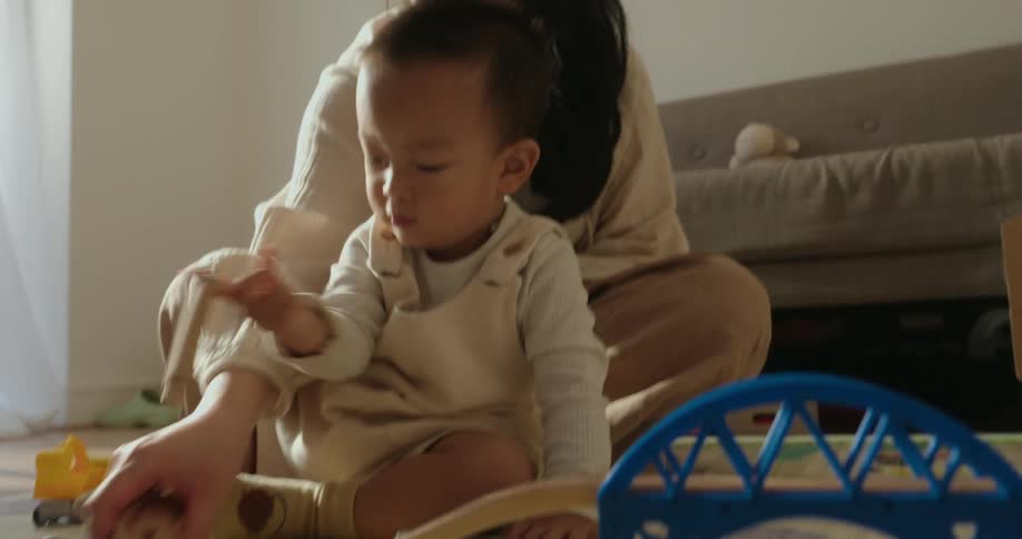 A mother and her baby are playing together on the floor in a sunlit living room surrounded by various toys the child wearing a white outfit engages with a wooden train track and a small wooden train car