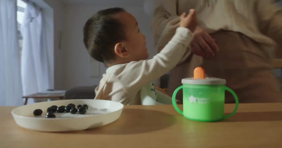 The video focuses on a toddler sitting at a table with a green sippy cup and a plate of blueberries in front of theman adults hands place the fruit down and the toddler then enthusiastically grabs a berry and feeds it to the adult who leans into the