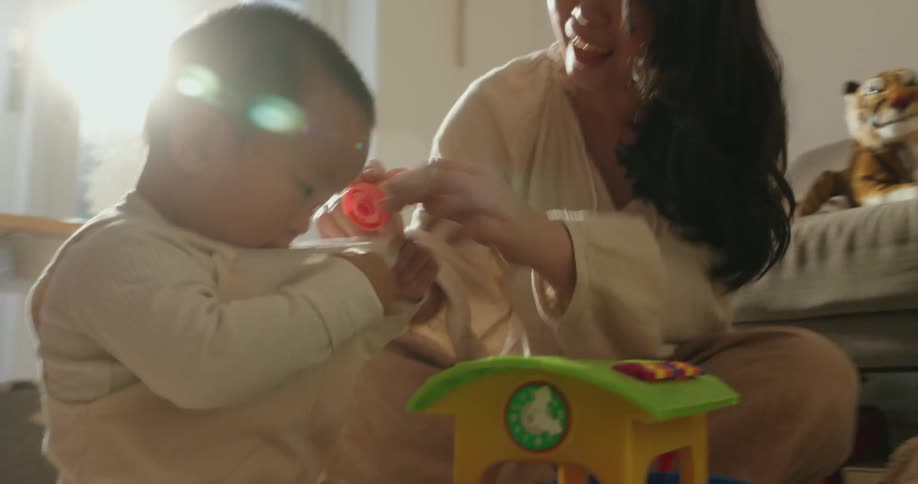 The video shows a warm closeup moment between a mother and her young child both sitting on the floor in a brightly lit roomthey are engaged with a colorful toy and the scene is filled with a soft bright light that creates a strong lens flare high