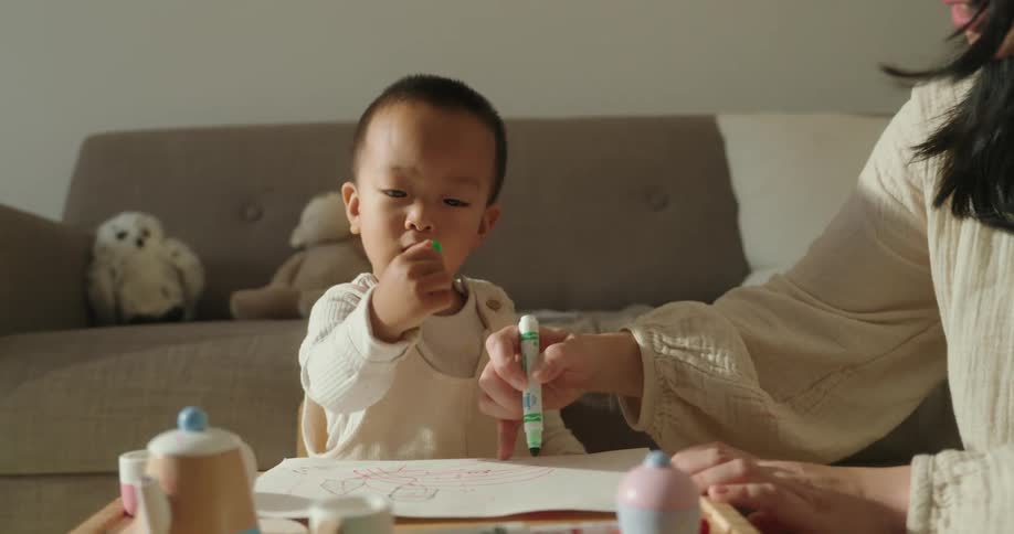 A little boy is playing with a green pen on a white sheet of paper and then hands it to an adult