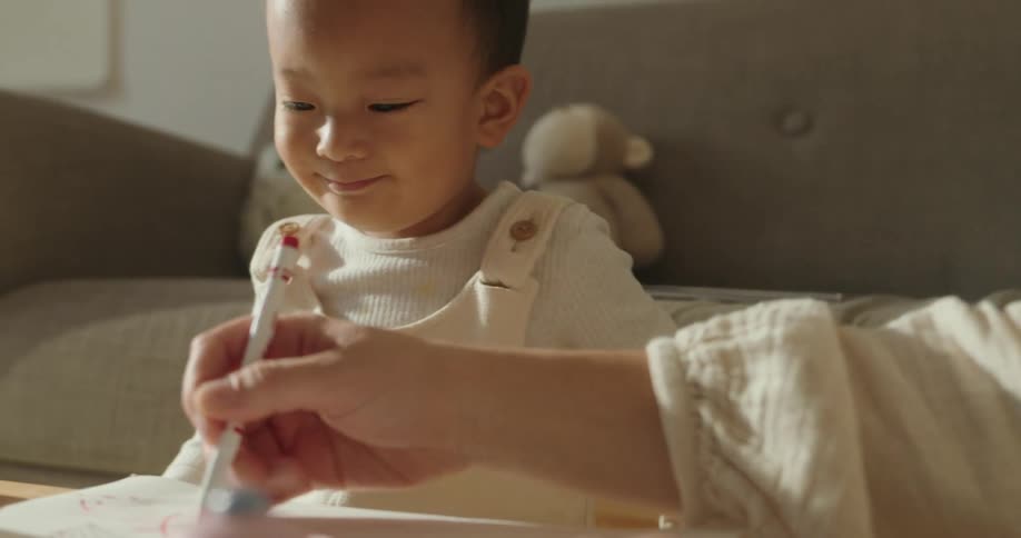 A toddler sits attentively watching an adults hand as they use a pen to draw or write on a sheet of paper the child smiles gently as they observe the activity with a teddy bear visible on the couch behind them