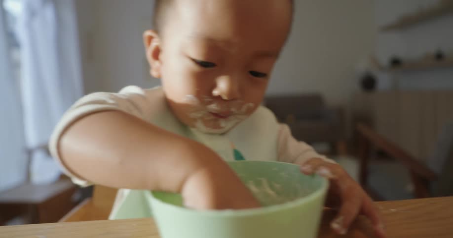 A toddler with food smeared around his mouth is focused on his meal digging his hands into a light green bowl he is wearing a white shirt and a bib while sitting in a high chair