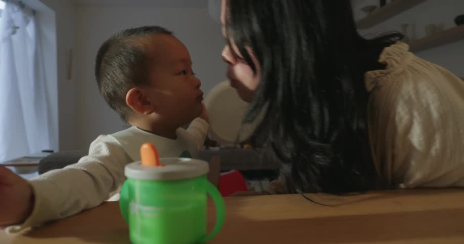 A woman with long dark hair leans in toward a baby seated in a high chair or at a table appearing to share a tender moment or a small kiss a bright green sippy cup is visible on the wooden table in the foreground of the indoor scene