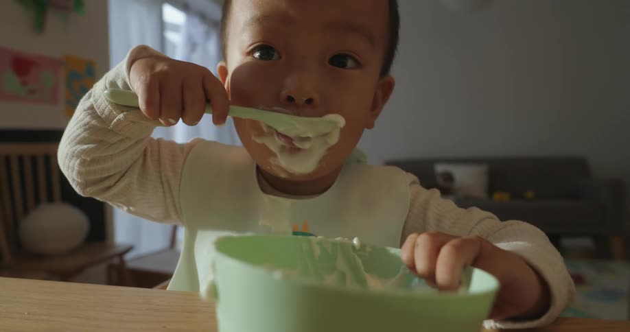 The video is a closeup shot of a toddler wearing a bib and sitting at a high chair or table fully engaged in selffeeding from a light green bowlthe child has food smeared all over their face and hands determinedly scooping a spoonful of white crea