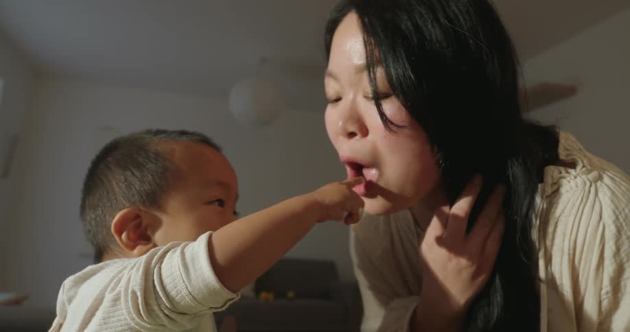 A child eating from a bowl with food stains on his face dipping his finger into the bowl to feed his mother at the dining table at midday