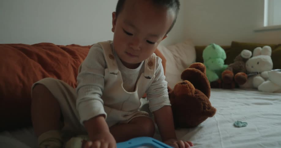 A baby sits on a bed playing with a small blue tray in a softly lit bedroom