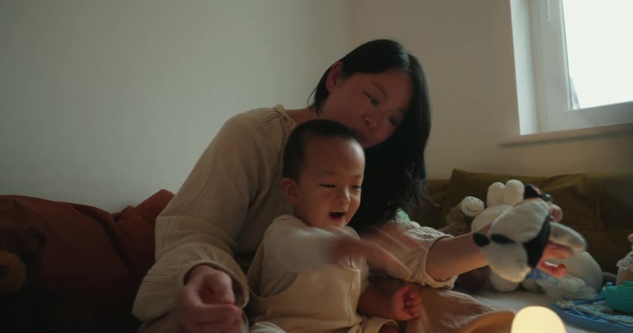 A mother playing with her baby son sitting on a bed in the bedroom surrounded by lots of toys