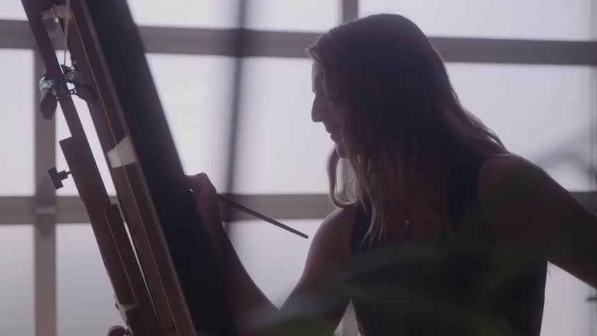 A young woman silhouetted against a large bright window is intensely focused on painting on a canvas supported by a wooden easel using a small paintbrush to apply detail to her artwork in an indoor studio setting