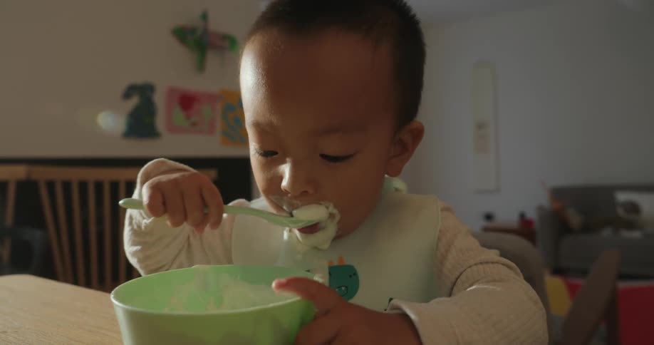 A toddler eats from a bowl with a spoon at a table in a softly lit home