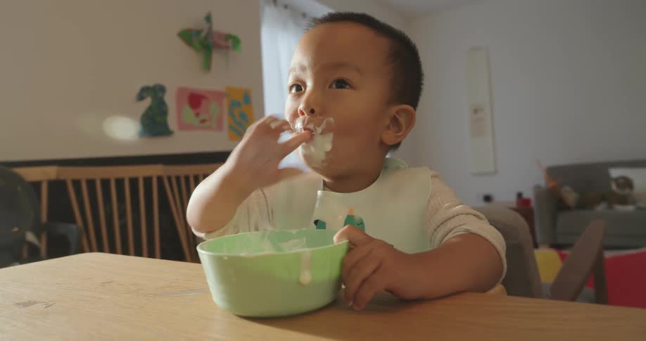 A little boy is eating white baby food from a green bowl and hes getting it all over himself