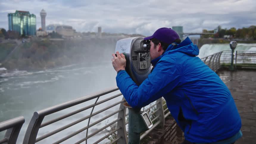 A young man wearing a blue jacket looks through a coinoperated binocular viewer at niagara falls at midday