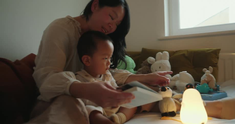 The video shows a warm intimate scene where a mother is sitting on a bed with her baby reading a book together they are surrounded by various stuffed animals and a glowing penguinshaped nightlight suggesting it is a bedtime or quiet time ritual