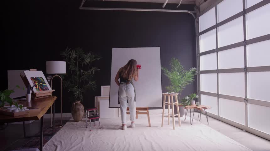 A woman in overalls paints a large canvas in a bright studio with plants and frosted garage doors