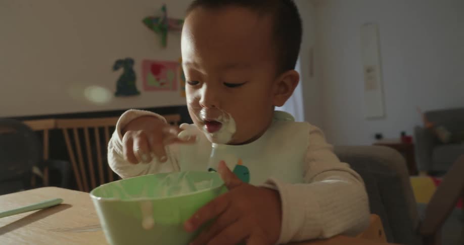 A toddler in a bib concentrates on scooping food from a bowl yogurt smeared around their mouth in a warmly lit home dining area
