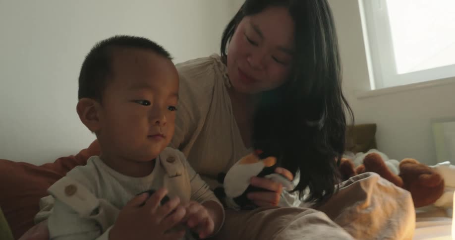A mother and son play with a stuffed toy on a bed in a softly lit bedroom