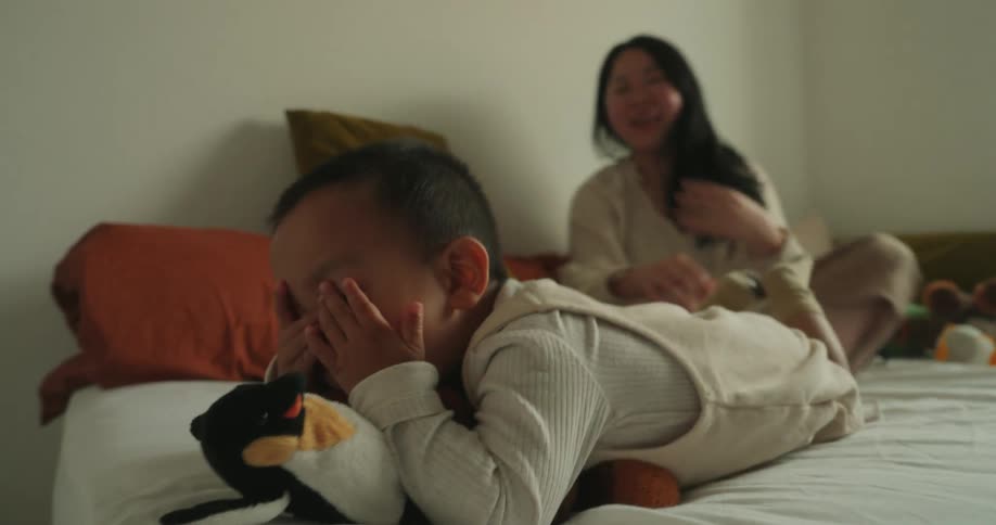 Child laughs while lying in bed holding a toy with her mother beside her