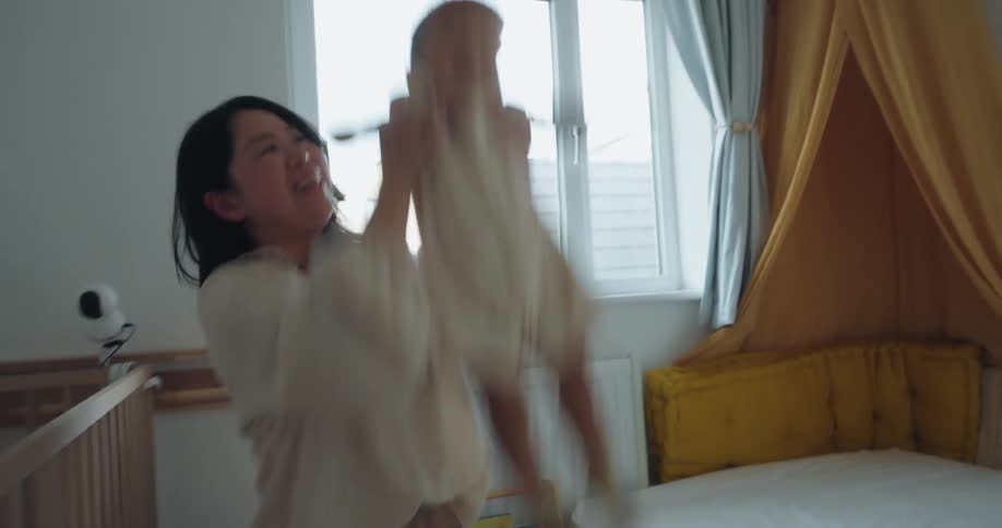 The video shows a joyful moment between a mother and her baby in a brightly lit nurserythe mother kneeling repeatedly lifts the giggling baby high into the air with the white window light streaming in behind them