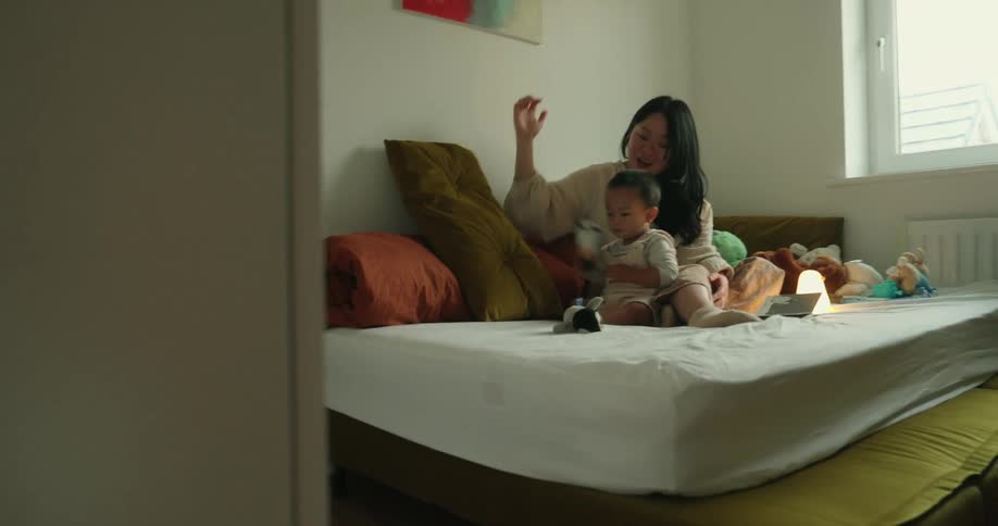 A mother and child sit on a bed playing with toys in a cozy sunlit bedroom