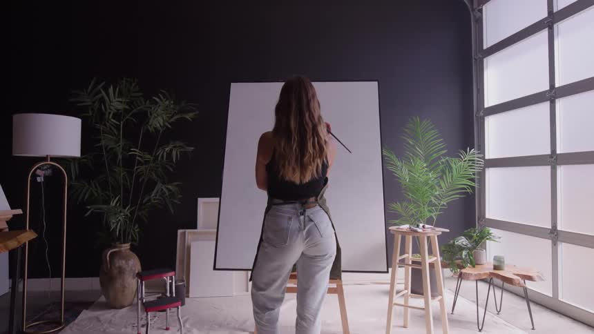 A woman stands at an easel painting on a large canvas in a bright studio