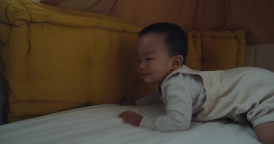 A toddler wearing a creamcolored outfit is seen crawling onto a bed and playfully burying their face into a large mustardyellow pillow on the headboard a woman then enters the frame leaning over to gently interact with the child