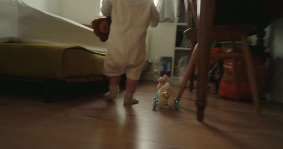 A small child wearing a lightcolored onesie and socks walks away from the camera clutching a brown teddy bear they are seen pulling a small toy on wheels across a wooden floor toward a bed in a sunlit room