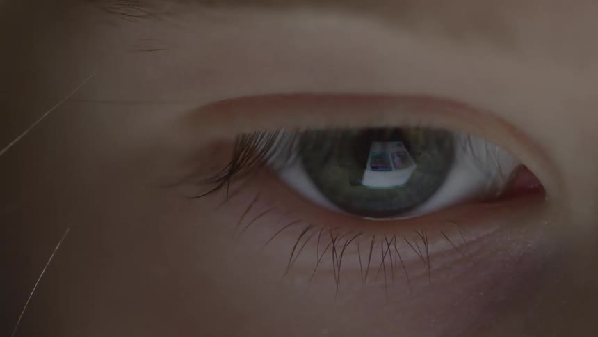 A closeup shot focuses on a partially open eye with a dark green iris and long eyelashes the reflection of a brightly lit monitor or screen can be clearly seen in the center of the pupil