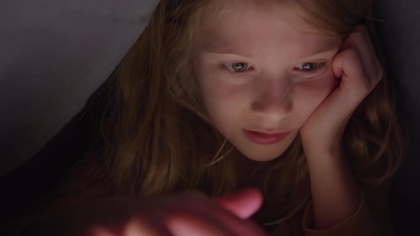 A young girl illuminated by the bright glow of a handheld device is captured in a closeup as her expression shifts from intense focus to a gentle smile while she interacts with the screes
